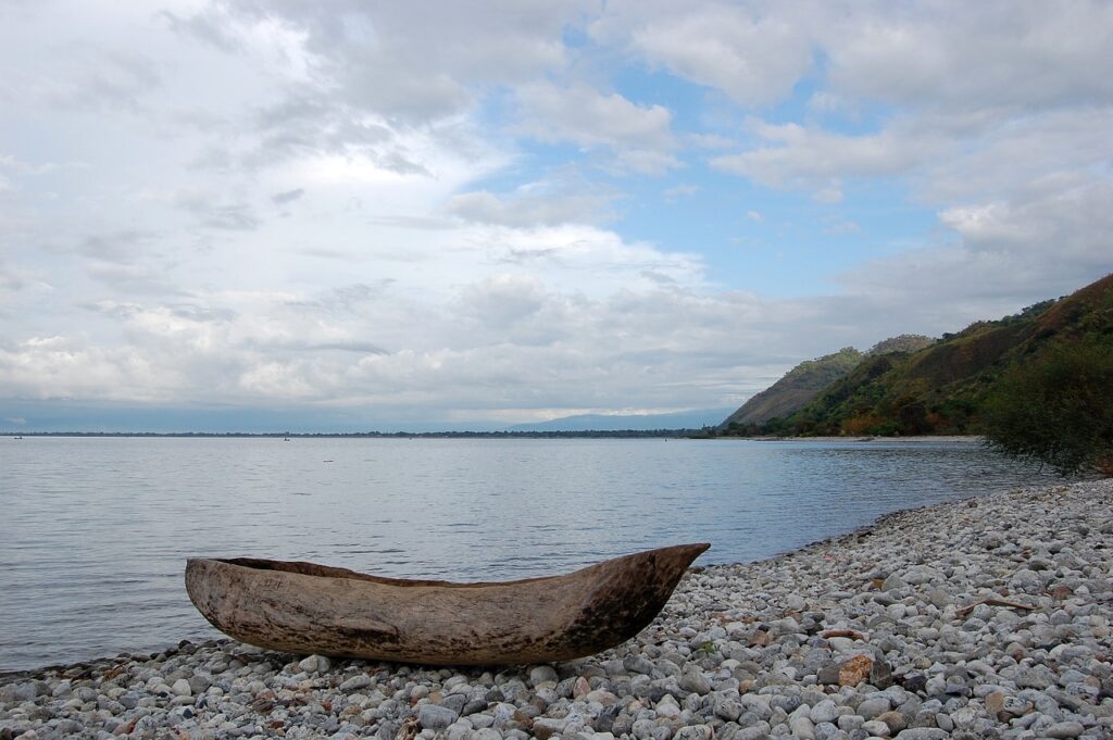 Shoreline of Lake Tanganyika with clear blue water, a dugout canoe, rocky outcrops, and lush greenery in the background.